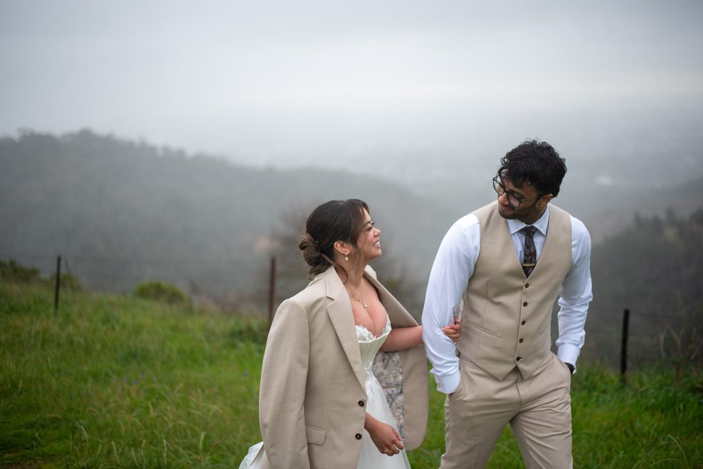 Bride and Groom walking through a field with the bride wearing the grooms jacket